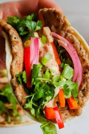 Someone holding a bahn mi smash burger taco with pickled veggies, sriracha mayo and cilantro.