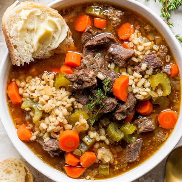 Top down shot of a bowl of beef and barley soup with a piece of bread sticking out of it.