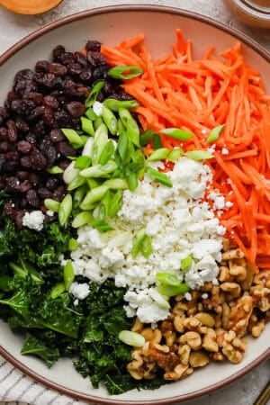 A bowl of carrot salad with raisins, kale, green onions, walnuts and goat cheese crumbles.