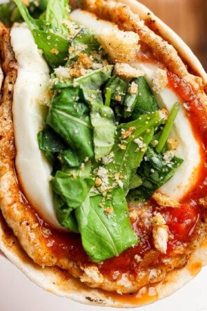 A hand holding a chicken parm smash taco that is topped with an arugula salad mixture and crushed breadcrumbs.