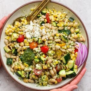 A big bowl of dense bean summer salad with two spoon sticking out and two hands holding the bowl.