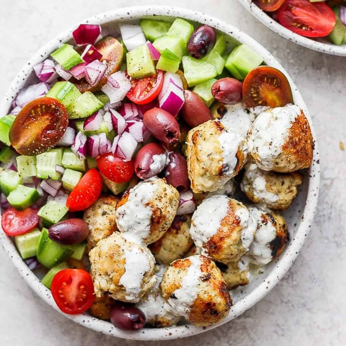 A bowl of greek chicken meatballs with tzatziki sauce and a greek cucumber salad.
