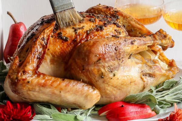 An Herbed Butter Turkey on a platter with red pears and herbs around it with someone basting the turkey with butter.