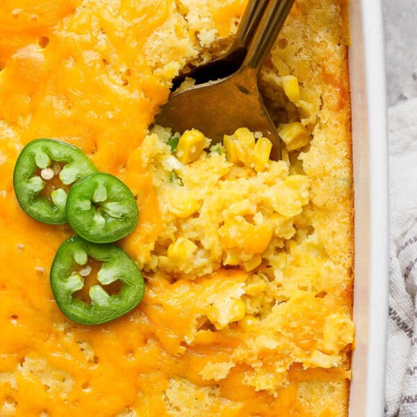 Top down, close up shot of a the corner of a pan of corn casserole with two spoons sticking out and three slices of jalapeno.