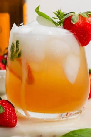A glass of a Kentucky Buck with ice inside and a strawberry next to it and sitting on the rim with a bottle of ginger beer in the background.