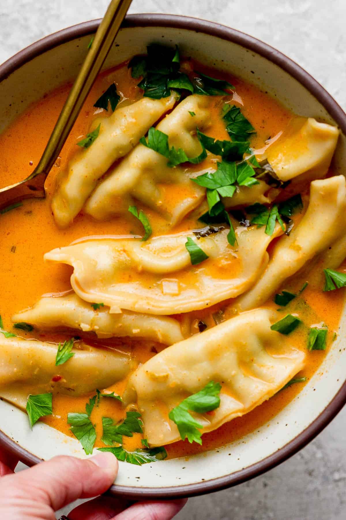 A hand holding a bowl of potsticker soup with a gold spoon sticking out the side.