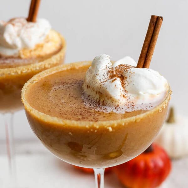 Two coupe glasses, each with a pumpkin pie martini in it, whipped cream and a cinnamon stick.