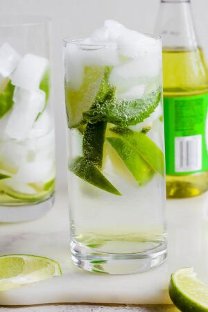 A glass of rose's lime juice gin and tonic with limes along with a bottle of rose's lime juice.