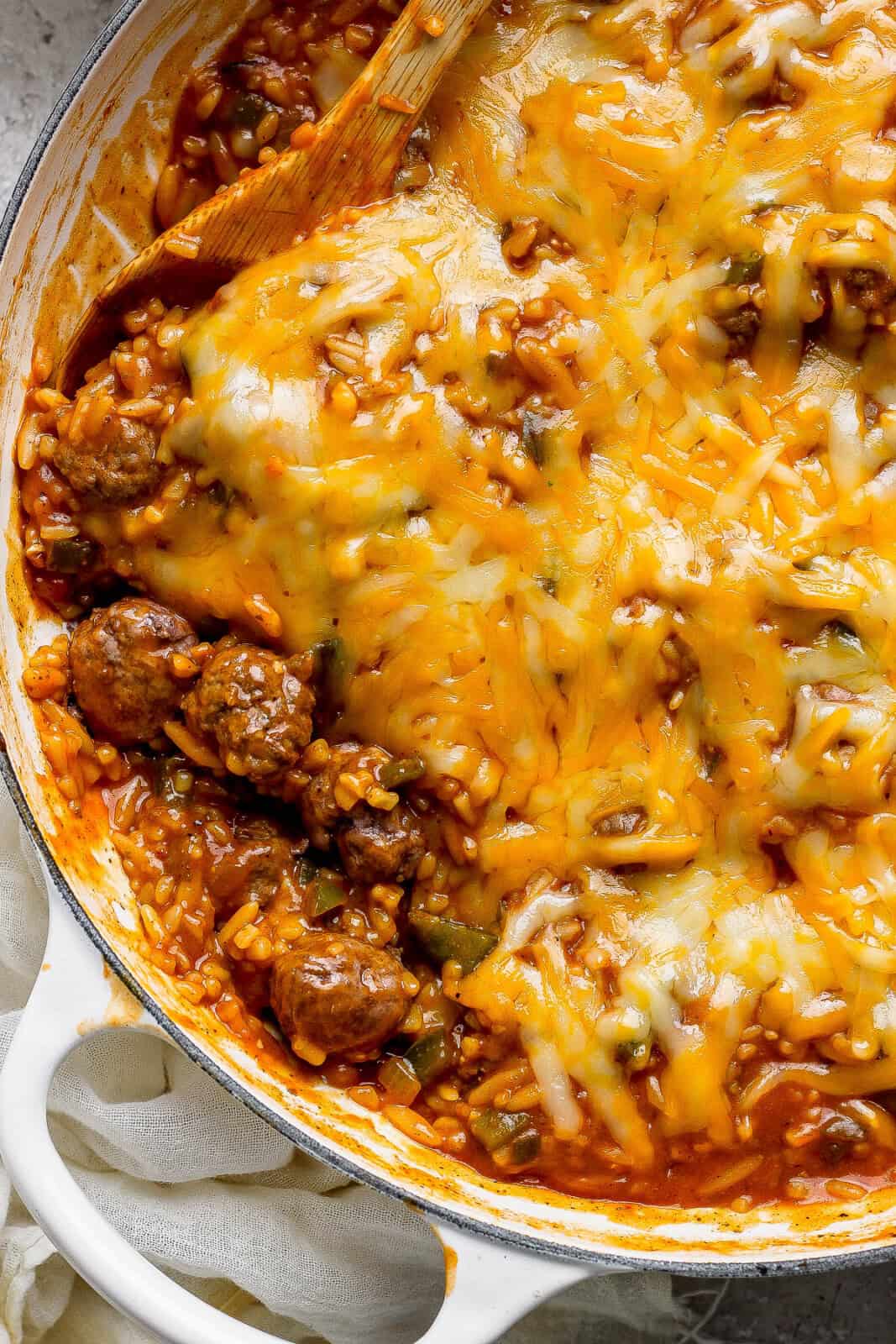 A wooden spoon in the side of sloppy joe orzo after it is fully cooked with cheese on top in a braising pan.