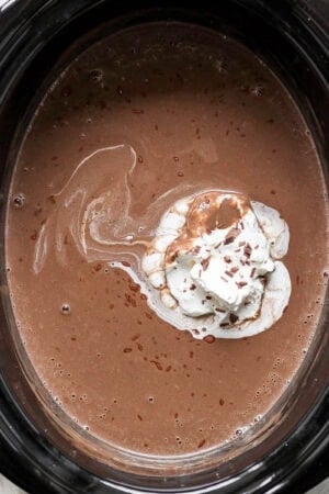 A crock pot fulled with homemade hot chocolate and whipped cream on top with shaved chocolate.