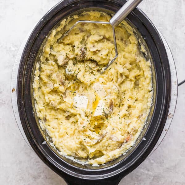 A large slow cooker filled with mashed potatoes and a potato masher sticking out.