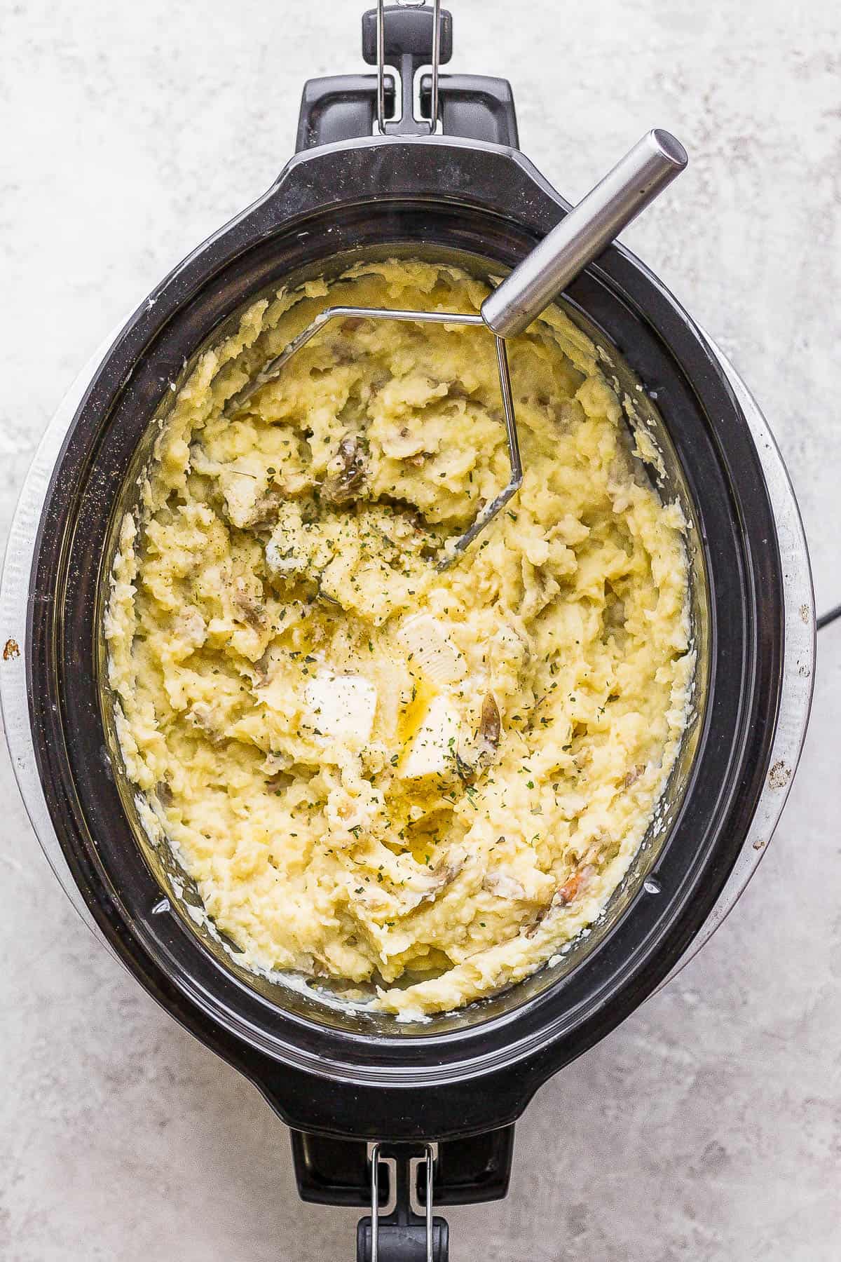 Slow cooker mashed potatoes in a slow cooker with a potato masher off to the side of the slow cooker. 
