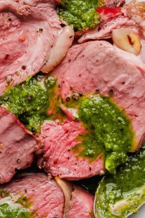 Sliced smoked leg of lamb on a plate with homemade chimichurri sauce over top and a spoon on the side.