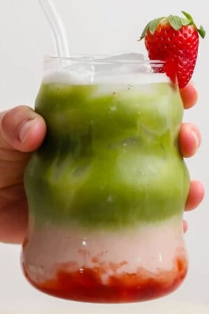 Someone holding a glass of strawberry match latte with a strawberry garnish and glass straw sticking out.