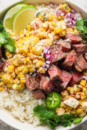 A bowl of rice topped with grilled steak and grilled street corn salad with lime wedges and fresh cilantro.