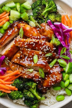 A teriyaki chicken rice bowl with grilled teriyaki chicken sliced over rice with carrots, grilled broccoli, edamame and green onions.