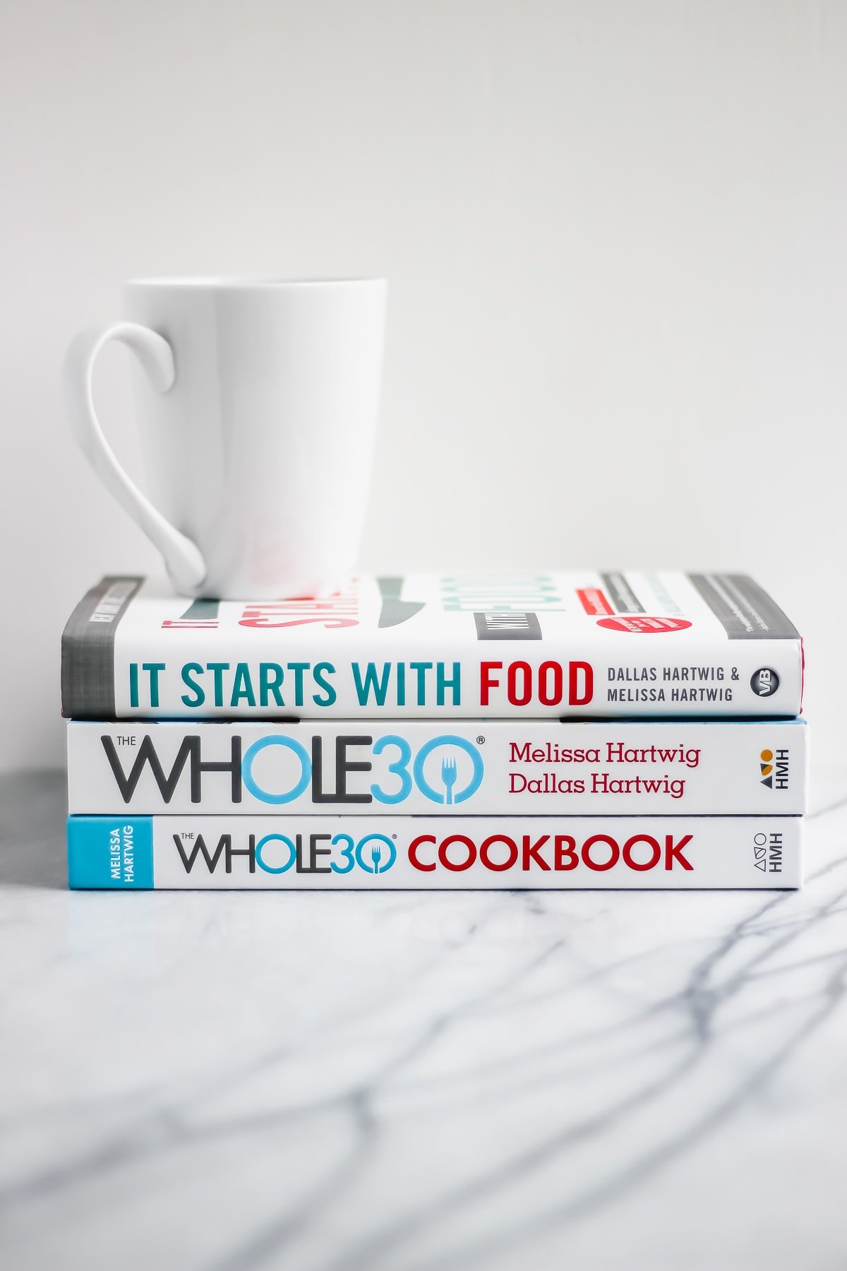 Three Whole30 books stacked on top of each other with a white coffee mug on top.